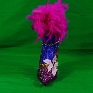 May include: A high-heeled shoe covered in blue and purple glitter, featuring a bright pink feather boa trim at the top, a gold chain accent around the ankle, and a single white and pink floral embellishment on the side of the pointed toe area.