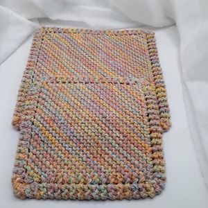 Peut inclure: Un gant de toilette rectangulaire tricoté à la main avec un motif de points en diagonale dans un mélange de fils variés aux tons pastel rose, jaune, bleu et blanc. Les bords sont finis par une bordure épaisse et texturée du même fil multicolore.