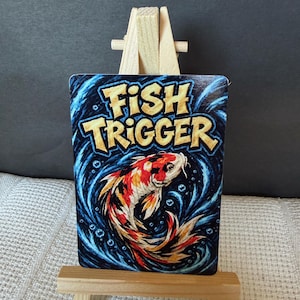Puede incluir: Una tarjeta rectangular con el texto FISH TRIGGER en letras amarillas llamativas sobre un pez koi colorido que nada en agua azul oscuro arremolinada, exhibida en un pequeño caballete de madera con tonos vibrantes de naranja, blanco y negro.