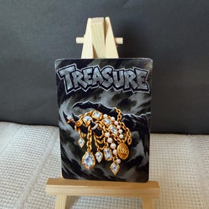 May include: A rectangular card featuring the word TREASURE in bold letters above an illustration of a dark, clawed hand holding gold chains with white pearls and clear, faceted crystals. The card is displayed on a small, light-coloured wooden easel.