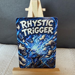 May include: A rectangular card featuring the text "RHYSTIC TRIGGER" in white letters at the top. The artwork depicts a profile of a figure with blue energy emanating from their head, surrounded by floating books in shades of blue and black.