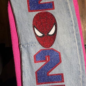 May include: Light blue denim jeans featuring a large red and blue glittery number 2027 design. A red and black glittery spider-themed mask is positioned between the numbers, and a black spider web graphic is printed near the top waistband.