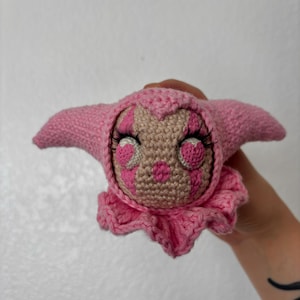 Pode incluir: Uma pequena boneca de crochê feita à mão com rosto castanho claro, marcas cor-de-rosa em forma de coração à volta dos olhos e nariz cor-de-rosa. A boneca usa um capuz cor-de-rosa pontiagudo e um colarinho com folhos, com pestanas longas.