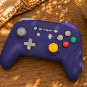 Könnte beinhalten: Ein violetter kabelloser Videospiel-Controller mit dem in Weiß aufgedruckten Text SWITCHOUSE. Das Gerät verfügt über ein graues Steuerkreuz, einen großen gelben Analogstick, einen kleineren grauen Analogstick sowie vier grüne, rote und graue Aktionstasten.
