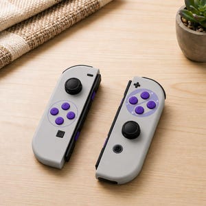 Pode incluir: Um par de controladores de videogame cinza-claros com botões de ação circulares roxos, joysticks pretos e trilhos laterais pretos. Os controladores têm um acabamento fosco liso e incluem um direcional na unidade direita e um botão quadrado na unidade esquerda.