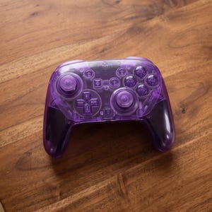 Può includere: Un controller per videogiochi wireless con un guscio in plastica viola traslucido. Il dispositivo include due levette analogiche, un pad direzionale, quattro pulsanti di azione principali e diversi pulsanti funzione sulla parte anteriore.