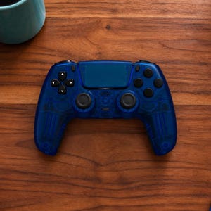 Op de afbeelding: Een doorschijnende blauwe draadloze videogamecontroller met zwarte analoge sticks, een richtingspad en actieknoppen. De controller heeft een centraal touchpad en interne componenten die zichtbaar zijn door de semi-transparante blauwe behuizing.