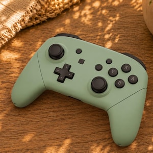 May include: A sage green wireless video game controller featuring black analogue sticks, a black directional pad, and four black action buttons labelled with the letters A, B, X, and Y. The controller has a matte finish and a central menu button.