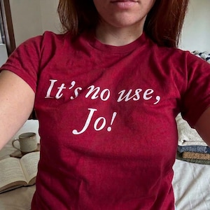May include: A maroon short-sleeved t-shirt featuring the white printed text "It's no use, Jo!" across the chest. The shirt has a crew neckline and is constructed from a solid-coloured fabric, providing a simple and direct design for casual wear.