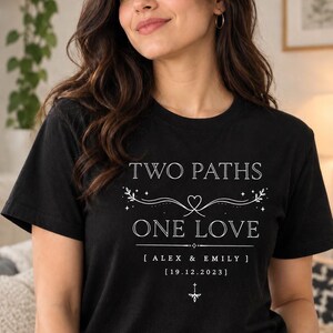 Könnte beinhalten: Ein schwarzes T-Shirt mit kurzen Ärmeln und weißem Aufdruck TWO PATHS ONE LOVE, den Namen ALEX & EMILY sowie dem Datum 19.12.2023. Das Design enthält dekorative Linien mit einem Herz, Sternen und floralen Elementen.