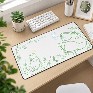 May include: A rectangular white desk mat featuring a green line art illustration of two frogs on a lily pad on the left and a larger frog blowing bubbles on the right, accented with thin green vines and a small yellow sparkle icon above the frogs.