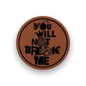 Puede incluir: Un posavasos circular marrón con un borde negro y el texto negro "YOU WILL NOT BREAK ME" en una fuente desgastada. El diseño incluye un icono de bomba, una corona, un círculo con patrón de rejilla y un símbolo de anarquía integrado.