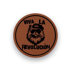 Könnte beinhalten: Ein runder Untersetzer aus braunem Leder mit einer schwarzen Grafik einer Katze, die ein Barett und eine Sonnenbrille trägt. Der Text VIVA LA REVOLUCION ist in Schwarz über und unter der Katzenillustration innerhalb eines schwarzen Randes gedruckt.