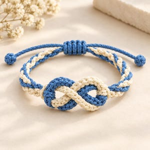 Puede incluir: Una pulsera de cordón trenzado con un diseño central de nudo infinito. La pulsera está construida con dos hebras entrelazadas de cuerda azul y color crema, con un cierre de nudo deslizante ajustable y pequeños nudos en los extremos de cada lado.