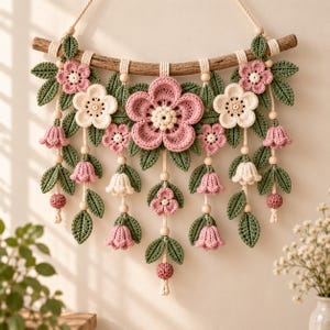 Puede incluir: Un adorno de pared con flores de ganchillo en tonos rosa y crema, hojas verdes y pequeñas cuentas redondas suspendidas de una rama de madera curvada. El arreglo incluye grandes flores centrales y flores pequeñas en forma de campana.