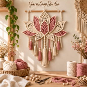 Könnte beinhalten: Ein Makramee-Wandbehang in Form einer Lotusblüte aus cremefarbenen und altrosa Kordeln, Holzperlen und Quasten, der an einem Holzstab hängt. Der Text YarnLoop Studio ist oben in der Mitte des Wandbehangs deutlich zu erkennen.