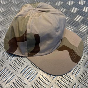 Peut inclure: Une casquette de baseball avec un motif camouflage désert dans des tons de beige, brun clair et brun foncé. La casquette présente une calotte structurée et une visière plate, fabriquée dans un tissu durable au tissage visible, avec six panneaux.
