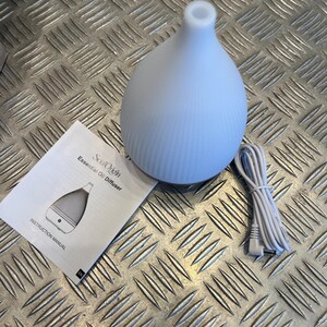 Peut inclure: Un diffuseur d'huiles essentielles blanc en forme de goutte avec une texture nervurée, accompagné d'un cordon d'alimentation blanc et d'un manuel d'instruction intitulé SoulOrigin Essential Oil Diffuser. L'appareil présente une ouverture supérieure arrondie.