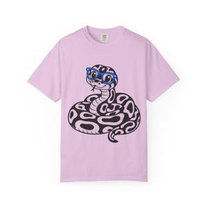 May include: A light purple short-sleeve t-shirt featuring a central graphic of a coiled snake with black and light purple patterned scales. The snake is wearing bright blue glasses and has a small black tongue visible near its mouth.