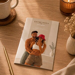 Peut inclure: Un livret rectangulaire intitulé ENTRE NOUS LE COUPLE présentant une photographie d'un couple prenant un selfie dans un miroir. La couverture affiche du texte en français et une image centrale de deux personnes s'enlaçant avec un smartphone.