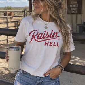 May include: A white short-sleeved t-shirt featuring the red text Raisin' HELL printed across the chest in a combination of script and block lettering. The shirt is paired with blue denim shorts, a brown and tan trucker hat, and a white insulated tumbler.