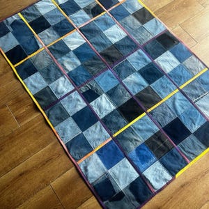 Op de afbeelding: Een rechthoekige patchwork quilt opgebouwd uit vierkante stukken spijkerstof in diverse blauwtinten. De quilt heeft een rasterpatroon met zichtbare naden en is afgewerkt met een felgele stoffen bies langs de randen voor contrast.
