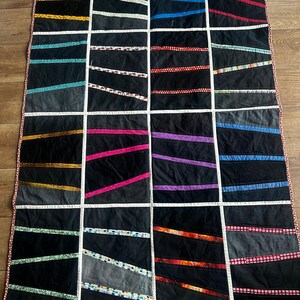 Könnte beinhalten: Ein rechteckiger Quilt mit einem Gitter aus schwarzen Stoffblöcken, die durch schmale weiße Streifen getrennt sind. Jeder Block enthält einen einzelnen diagonalen Streifen aus gemustertem oder einfarbigem Stoff in verschiedenen Farben wie Gelb, Blau, Rot und Grün.