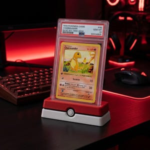 Peut inclure: Une carte à collectionner Pokemon Charmander de 1999, classée PSA numéro 46 avec une note GEM MT 10. La carte est enfermée dans un étui en plastique transparent et présentée sur un support rouge et blanc sur le thème d'une Pokeball.