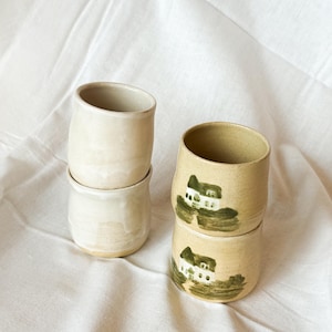 Peut inclure: Deux piles de tasses en céramique. La pile de gauche se compose de deux tasses unies, blanc cassé, à la surface lisse. La pile de droite présente deux tasses beige clair mouchetées, ornées chacune d'une illustration de maison peinte en vert et blanc.