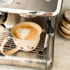 Puede incluir: Una taza de cerámica moteada con un patrón de ovejas pintado a mano descansa sobre la bandeja de goteo de una máquina de café espresso de acero inoxidable. La taza contiene un latte con arte en espuma. La máquina muestra el texto Ultima Cosa.