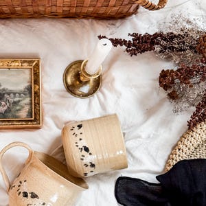 Peut inclure: Deux tasses en céramique beige avec des motifs noirs tachetés, un bougeoir en laiton avec une bougie blanche, un petit cadre photo, un panier en osier, des fleurs séchées et un morceau de tissu noir sur une surface en tissu blanc.