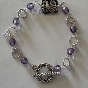 May include: A bracelet featuring purple faceted beads and silver-toned metal links. The piece includes a square charm embossed with a bird and flower design, and a circular toggle clasp with a textured edge and a leaf-shaped bar closure.
