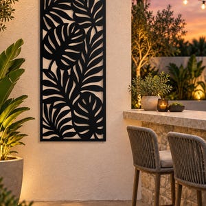 May include: A rectangular black metal wall panel featuring a laser-cut design of overlapping tropical monstera and palm leaves. The panel is mounted vertically on a light-coloured textured wall, displaying intricate botanical silhouettes.