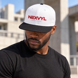 Pode incluir: Um boné de beisebol branco com aba preta e um botão superior preto. A parte frontal do boné apresenta o texto vermelho NEXVYL com as palavras next level impressas em letras vermelhas menores diretamente abaixo do nome da marca.