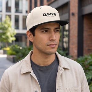 May include: A cream-coloured baseball cap with a black brim and the black text QAVYR printed on the front panel. The cap features a structured crown and a flat brim design, providing a clean and simple look for casual headwear accessories.