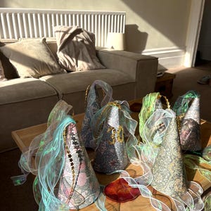 May include: A collection of four cone-shaped party hats featuring a detailed floral pattern in shades of blue, green, and pink. Each hat is adorned with iridescent, translucent ribbon streamers and a thin gold string with small star accents.