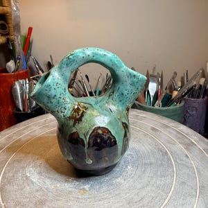 May include: A ceramic vessel with a rounded base and two upward-pointing spouts connected by a central arched handle. The piece features a glossy glaze with a mottled turquoise and green top section that transitions into a dark brown base.