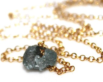 May include: A delicate gold chain necklace with a raw, gray-green diamond pendant. The diamond is rough and unpolished, giving it a natural and unique look.