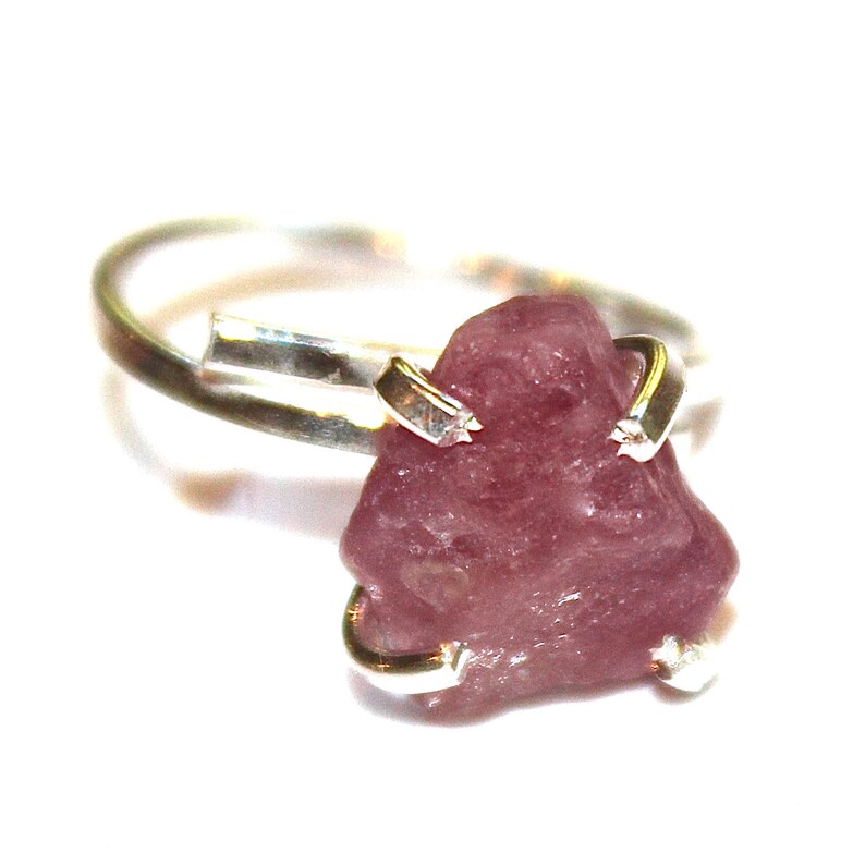 May include: A silver ring featuring a raw pink gemstone set in the centre. The gemstone is rough, with an irregular shape and a natural, unpolished finish.