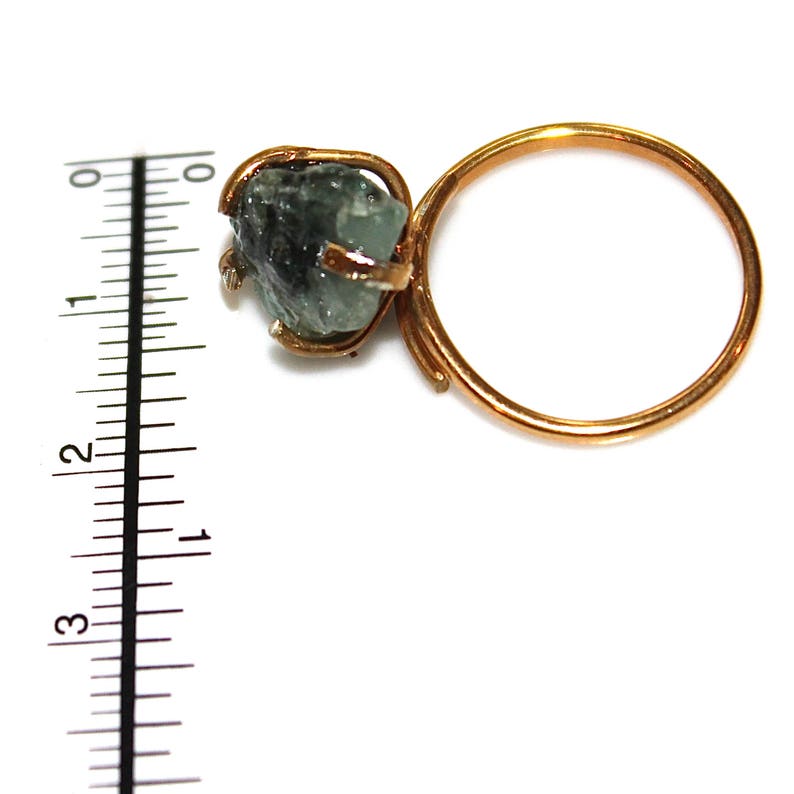 Pu&ograve; includere: Un anello d'oro con una pietra preziosa verde incastonata in una griffa. L'anello &egrave; su una superficie bianca con un righello sullo sfondo.