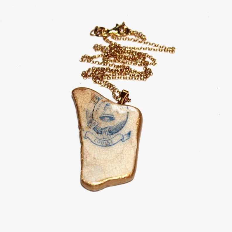 May include: A gold chain necklace with a pendant made from a white ceramic shard with a blue and white design. The design features a bell, a banner with the word "LOUISE", and other decorative elements.