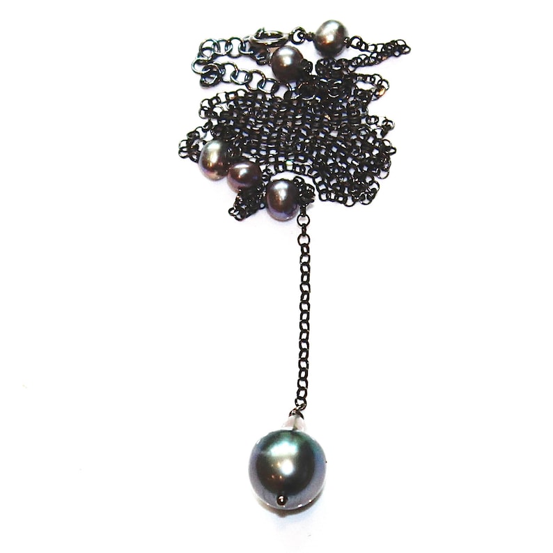 May include: A black chain necklace with a long drop featuring a large, dark green pearl. The necklace is adorned with several smaller pearls.