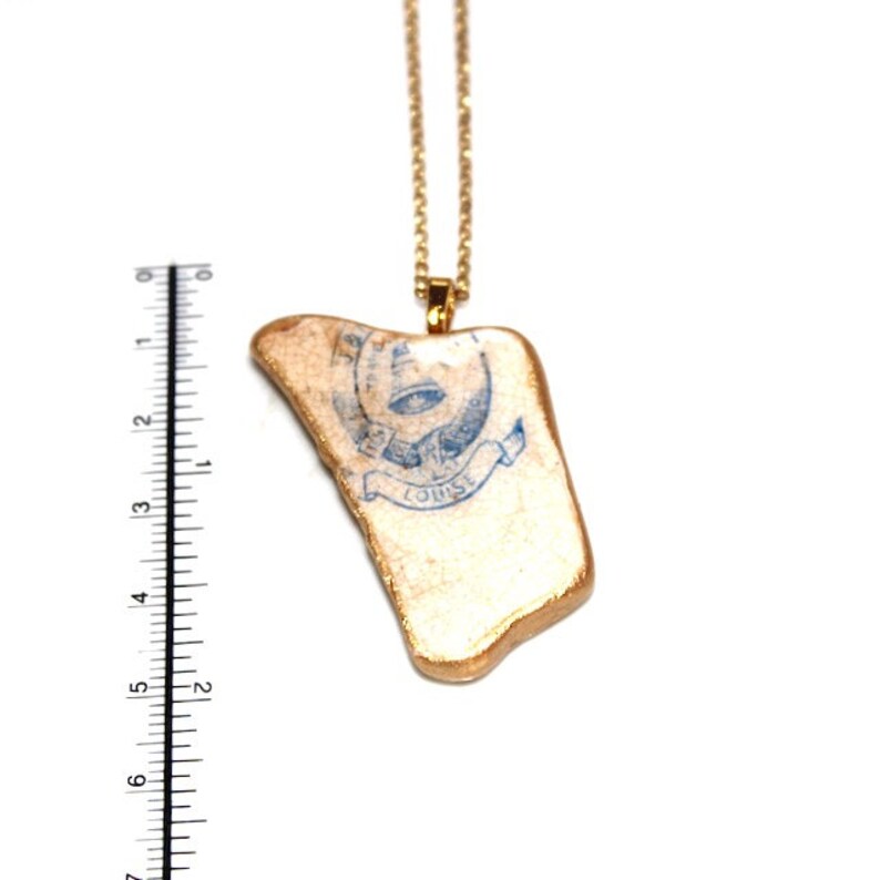 May include: A gold chain necklace with a pendant made from a piece of ceramic with a blue and white design. The design features an eye with a banner that reads "Louise".