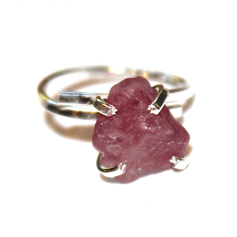 May include: A silver ring with a raw pink gemstone set in the centre. The gemstone is held in place by silver prongs.