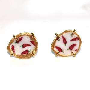 May include: A pair of gold-toned earrings with a white background and red chili pepper pattern. The earrings are made of metal and have a unique, irregular shape.