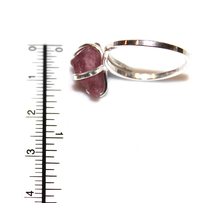 May include: A silver ring with a pink crystal stone set in a wire wrap. The ring is shown next to a ruler, with the stone measuring approximately 2.5 cm in length.