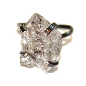 May include: A clear quartz crystal ring with a black metal band. The crystal is faceted and has a rough, natural look.