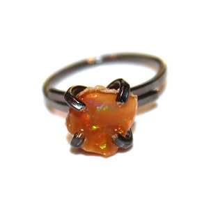 Könnte beinhalten: Ein Ring mit einem großen, orangefarbenen Opal-Edelstein, der in eine schwarze Metallfassung gefasst ist. Der Opal zeigt einen Regenbogen an Farben.