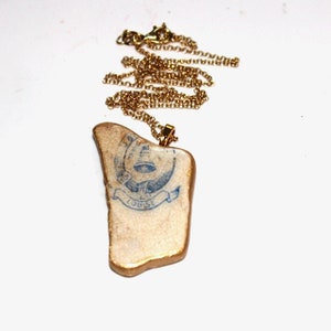 May include: A gold chain necklace with a pendant made from a piece of ceramic with a blue and white design. The design features an eye, a bell, and the word "HOUSE".