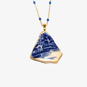May include: A gold chain necklace with a blue and white porcelain pendant. The pendant is a shard of a broken plate with a gold rim. The porcelain features a blue and white landscape design.